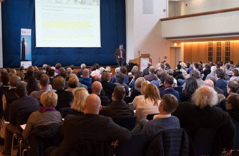 Publikum in einem Saal blickt auf eine Präsentationsleinwand
